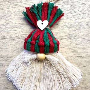 Może przedstawiać: Ręcznie robiona ozdoba świąteczna z krasnalem. Krasnal ma czerwono-zieloną czapkę, białą brodę i drewniany nos. Do czapki przymocowane jest białe serce. Ozdoba zawieszona jest na sznurku jutowym.