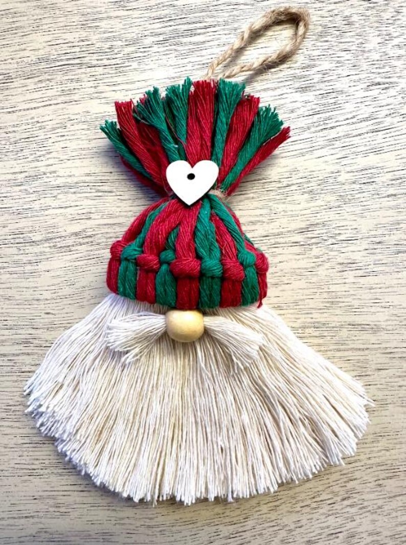 Może przedstawiać: Ręcznie robiona ozdoba świąteczna z krasnalem. Krasnal ma czerwono-zieloną czapkę, białą brodę i drewniany nos. Do czapki przymocowane jest białe serce. Ozdoba zawieszona jest na sznurku jutowym.
