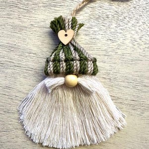 Może przedstawiać: Ręcznie robiona ozdoba gnoma z zielono-szaro-białą dzianinową czapką, drewnianym sercem i długą białą brodą z frędzli. Drewniana kulka służy jako nos, a sznurek jutowy jest przymocowany do zawieszenia.