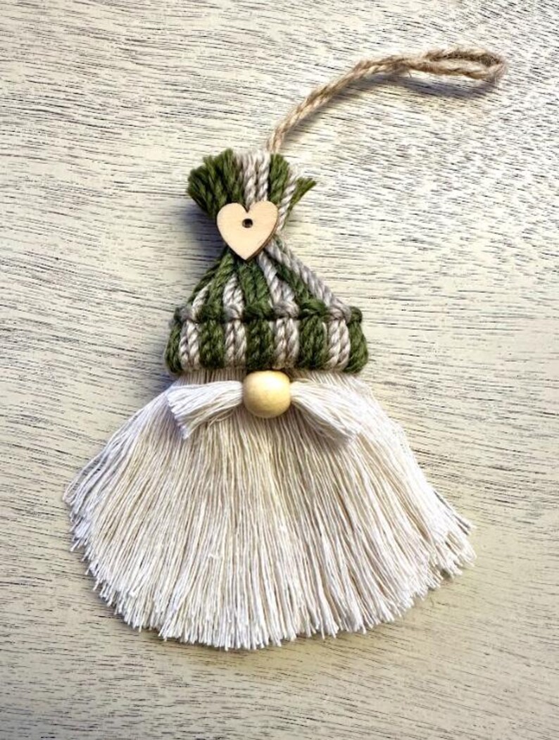 Może przedstawiać: Ręcznie robiona ozdoba gnoma z zielono-szaro-białą dzianinową czapką, drewnianym sercem i długą białą brodą z frędzli. Drewniana kulka służy jako nos, a sznurek jutowy jest przymocowany do zawieszenia.