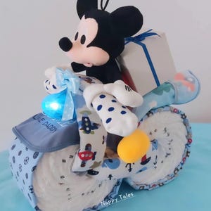 May include: A blue and white nappy cake shaped like a motorbike with a Mickey Mouse plush toy riding on top. The motorbike has a blue light on the front and a gift box tied with a blue ribbon on the back. The cake is decorated with a blue and white polka dot pattern and the words "Like Daddy" on the side. The cake is sitting on a blue surface.