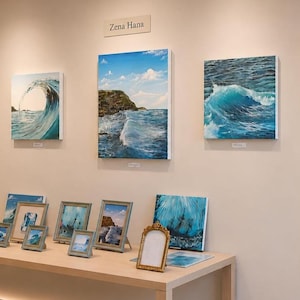 May include: A gallery display featuring multiple framed and unframed canvas prints of ocean waves and coastal landscapes. The wall art showcases various shades of blue and green water, with a sign reading Zena Hans centered above the collection.
