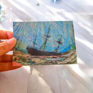 May include: A rectangular sticker featuring a dark brown sailing ship resting on the ocean floor. The scene includes blue water, coral, and a holographic finish that creates a shimmering effect of small stars across the entire surface.