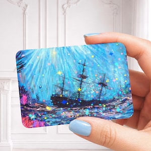 May include: A rectangular card featuring a vibrant underwater scene with a dark silhouette of a sailing ship. The image is overlaid with a holographic star pattern that reflects light in various colors against the deep blue ocean background.