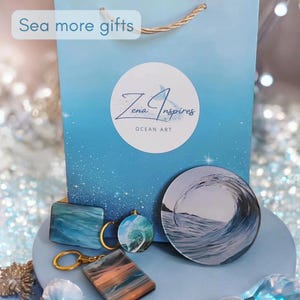 May include: A light blue gift bag with the text "Sea more gifts" and a circular logo reading "Zena Inspires OCEAN ART". In front are two rectangular keychains with blue and orange wave patterns, a circular wave keychain, and a round wave coaster.