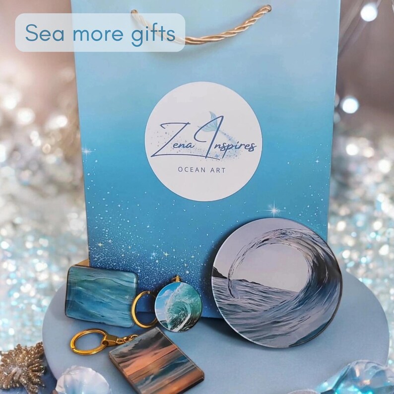 May include: A light blue gift bag with the text "Sea more gifts" and a circular logo reading "Zena Inspires OCEAN ART". In front are two rectangular keychains with blue and orange wave patterns, a circular wave keychain, and a round wave coaster.