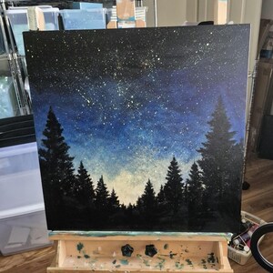 May include: A square canvas painting depicting a night sky filled with white and yellow star-like speckles above a dark silhouette of a dense pine forest. The sky transitions from a deep black at the top to a lighter blue and white near the horizon.