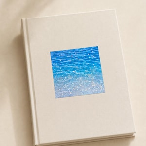 K&ouml;nnte beinhalten: Ein geschlossenes, cremewei&szlig;es Hardcover-Buch mit einem quadratischen Aufkleber auf dem Vorderdeckel. Der Aufkleber zeigt eine leuchtend blaue Wasseroberfl&auml;che mit schimmernden Lichtreflexen, die einen hellen, klaren und aquatischen Effekt erzeugen.