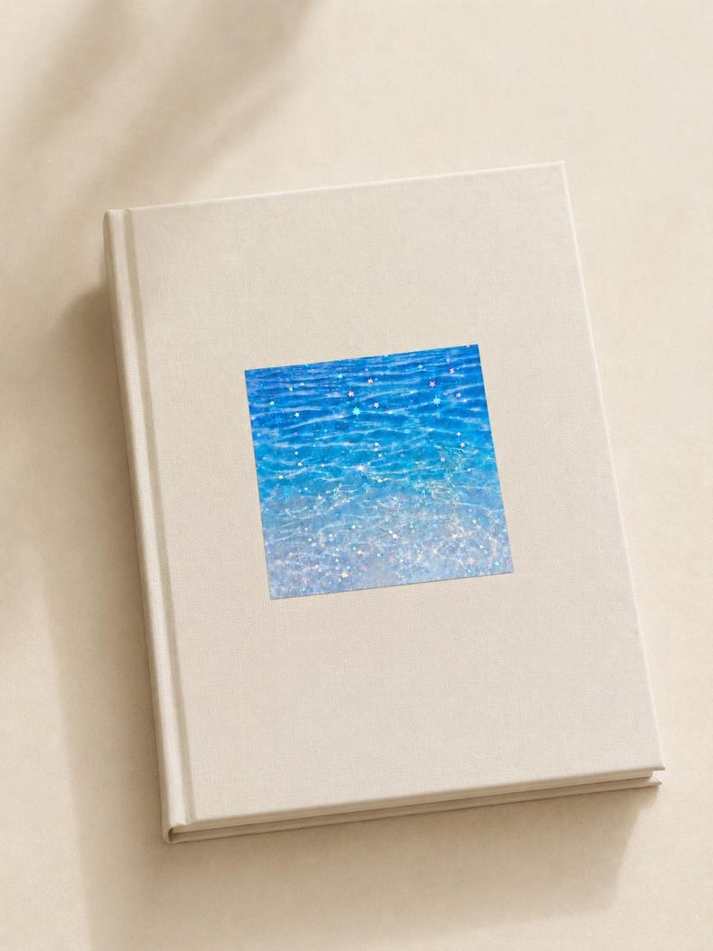 K&ouml;nnte beinhalten: Ein geschlossenes, cremewei&szlig;es Hardcover-Buch mit einem quadratischen Aufkleber auf dem Vorderdeckel. Der Aufkleber zeigt eine leuchtend blaue Wasseroberfl&auml;che mit schimmernden Lichtreflexen, die einen hellen, klaren und aquatischen Effekt erzeugen.