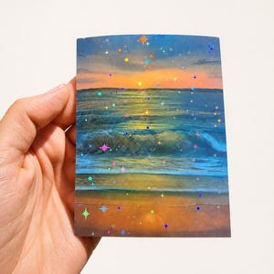 May include: A rectangular print featuring a sunset over the ocean with waves rolling onto a sandy beach. The image is overlaid with a holographic star pattern that reflects light in various colors, including blue, pink, yellow, and orange.