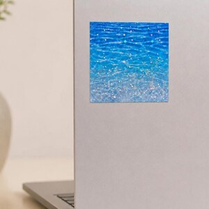 K&ouml;nnte beinhalten: Ein quadratischer Aufkleber mit einer leuchtend blauen Wasseroberfl&auml;che, die einen Farbverlauf von tiefem Marineblau bis zu hellem Cyan aufweist. Das Design ist mit holografischen Sternen &uuml;berlagert, die schimmernde Lichtreflexe erzeugen.