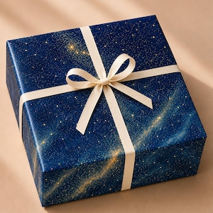 May include: A square gift box wrapped in dark blue paper featuring a gold celestial galaxy print with stars and nebulae. The box is tied with a smooth, cream-colored ribbon that forms a centered bow on the top surface of the package.