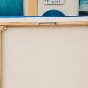 May include: The back of a rectangular canvas stretched over a light-colored wooden frame. The canvas is secured with staples along the edges and features a silver-toned metal sawtooth hanger centered on the top wooden bar for wall mounting.
