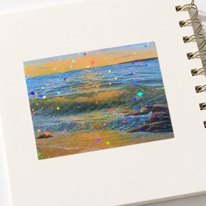 May include: A rectangular sticker featuring a coastal sunset scene with ocean waves, sandy shores, and dark rocks, overlaid with a holographic star pattern that reflects various colors across the blue, orange, and yellow landscape.