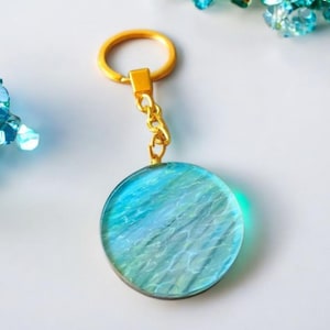 May include: A round keychain with a gold-colored ring and chain. The pendant is a translucent circle with blue and green hues, resembling water. The keychain is on a white surface.