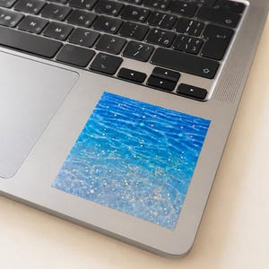 K&ouml;nnte beinhalten: Ein quadratischer Aufkleber mit einer leuchtend blauen Wasseroberfl&auml;che mit Wellen und Lichtreflexionen, &uuml;berlagert mit einem Muster aus kleinen, mehrfarbigen holografischen Sternen, die &uuml;ber das gesamte Design verteilt sind, auf einem silbernen Laptop.