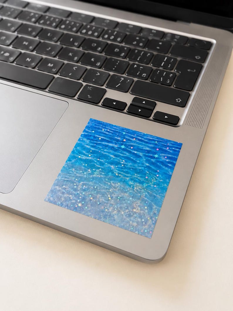 K&ouml;nnte beinhalten: Ein quadratischer Aufkleber mit einer leuchtend blauen Wasseroberfl&auml;che mit Wellen und Lichtreflexionen, &uuml;berlagert mit einem Muster aus kleinen, mehrfarbigen holografischen Sternen, die &uuml;ber das gesamte Design verteilt sind, auf einem silbernen Laptop.