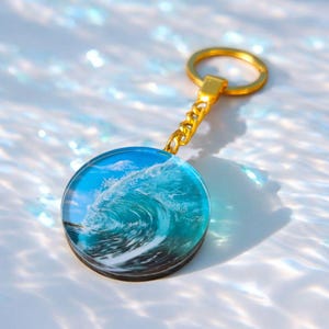 May include: A round keychain featuring a detailed ocean wave design in shades of blue and white. The wave is encased in clear resin, attached to a gold-colored chain and keyring. The keychain is a decorative accessory.