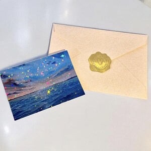 May include: A rectangular postcard featuring a seascape with a dark blue ocean and a cloudy sky, overlaid with a holographic star pattern. Beside it lies a light beige envelope sealed with a circular gold wax seal embossed with a letter V.