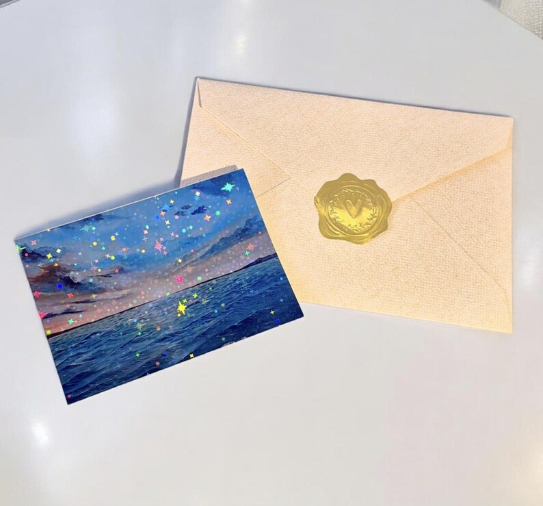 May include: A rectangular postcard featuring a seascape with a dark blue ocean and a cloudy sky, overlaid with a holographic star pattern. Beside it lies a light beige envelope sealed with a circular gold wax seal embossed with a letter V.