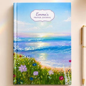 Pode incluir: Um diário de capa dura com uma pintura a óleo de uma paisagem costeira com um oceano iluminado pelo sol, ondas a quebrar e um primeiro plano de flores silvestres vibrantes em tons de roxo, vermelho e branco. O texto Emma's PRAYER JOURNAL está centrado.