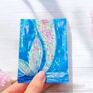 May include: A rectangular sticker featuring a mermaid tail design. The tail is iridescent with shades of pink, purple, and silver, set against a blue background with star accents. The mermaid tail is in the center of the sticker.