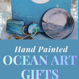 May include: A collection of ocean-themed items including a circular keychain with a wave design, a blue gift bag with the text Zena Inspires OCEAN ART, and a rectangular card featuring a holographic mermaid tail illustration with star accents.