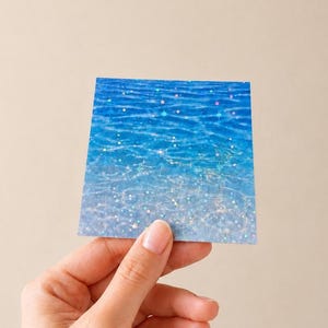 K&ouml;nnte beinhalten: Ein quadratischer Aufkleber mit einem leuchtend blauen Wasseroberfl&auml;chenmuster und holografischem Finish. Das Design zeigt kr&auml;uselnde Wellen in Azur- und Cyan-T&ouml;nen, die mit einer Streuung kleiner, schimmernder sternf&ouml;rmiger Symbole &uuml;berlagert sind.