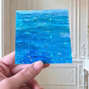 May include: A square sticker featuring a vibrant blue water surface pattern with a holographic finish. The design is covered in numerous small, multi-pointed star shapes that shimmer with iridescent light across the deep blue aquatic waves.
