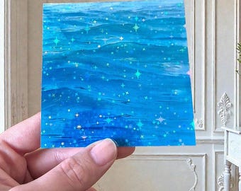 Könnte beinhalten: Ein quadratischer Aufkleber mit einem leuchtend blauen Wasseroberflächenmuster und holografischer Oberfläche. Das Design ist mit zahlreichen kleinen, mehrzackigen Sternen bedeckt, die in schillerndem Licht über den tiefblauen Wellen funkeln.