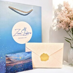 May include: A blue gift bag with gold rope handles featuring a white circular logo that reads Zena Inspires OCEAN ART, a small rectangular card with a sunset ocean scene, and a light peach envelope sealed with a circular gold wax seal.