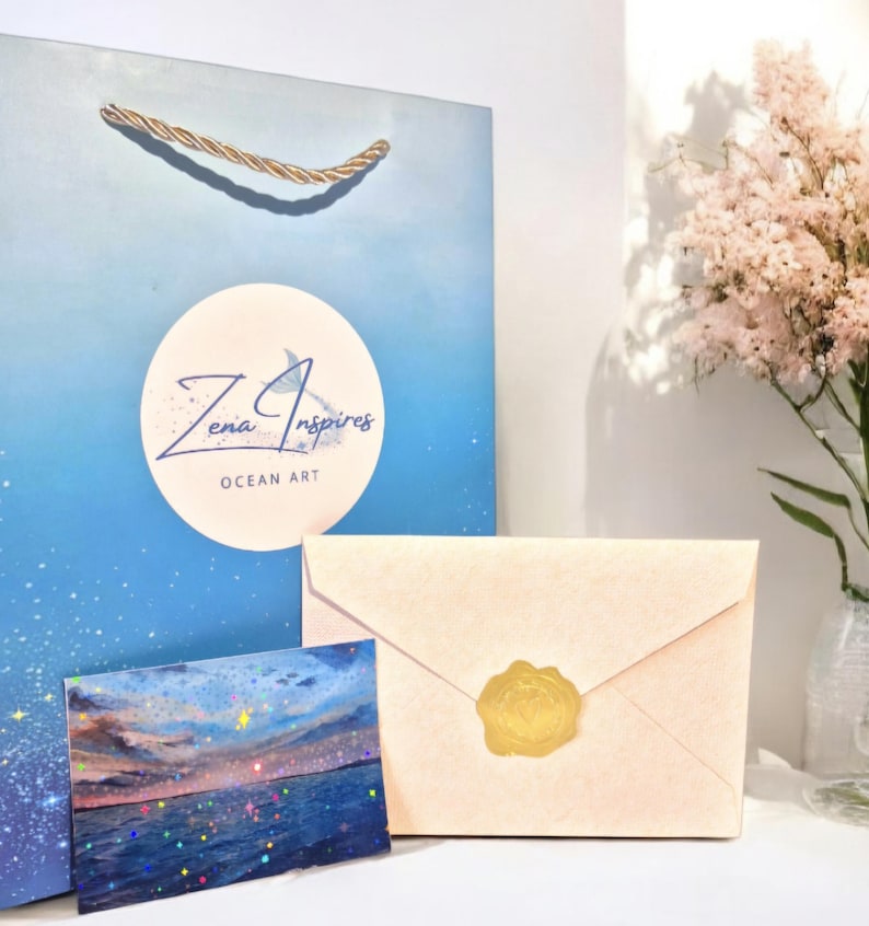 May include: A blue gift bag with gold rope handles featuring a white circular logo that reads Zena Inspires OCEAN ART, a small rectangular card with a sunset ocean scene, and a light peach envelope sealed with a circular gold wax seal.