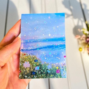 May include: A rectangular sticker featuring a landscape illustration of a blue ocean under a light blue sky, with a foreground of colorful wildflowers. The surface has a holographic finish with small star-shaped sparkles scattered throughout.