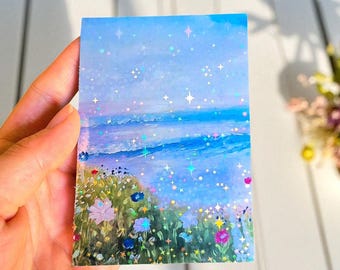 Könnte beinhalten: Ein rechteckiger Aufkleber mit einer Landschaftsillustration, die einen blauen Ozean unter einem hellblauen Himmel und bunte Wildblumen im Vordergrund zeigt. Die Oberfläche hat ein holografisches Finish mit kleinen sternförmigen Glitzereffekten.