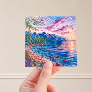 May include: A square art print featuring a colorful landscape painting of a lake at sunset. The scene includes deep blue water with scattered rocks, a shoreline with green trees, and a vibrant sky filled with pink and purple clouds above mountains.