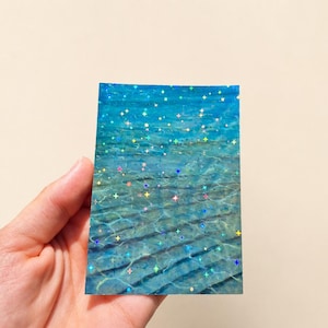May include: A rectangular card featuring a photographic print of clear blue water with rippling light patterns on the sandy bottom, overlaid with a holographic finish that displays small, multi-colored, four-pointed star shapes.