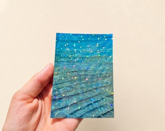 Könnte beinhalten: Eine rechteckige Karte mit einem Fotodruck von klarem blauem Wasser mit wellenförmigen Lichtmustern auf dem sandigen Boden, überzogen mit einem holografischen Finish, das kleine, mehrfarbige, vierzackige Sternformen zeigt.