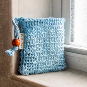 May include: A light blue, open-weave soap bag with a drawstring closure and a wooden bead. The bag is made of a woven material and has a small tag that reads "Pacific Glow". The bag is sitting on a windowsill.