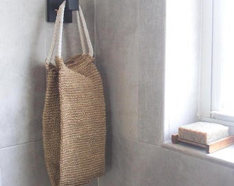 May include: A rectangular woven tan jute storage bag with two thick white braided rope handles hanging from a square black wall hook. The bag features a textured, natural fiber construction and is suspended vertically against a tiled wall surface.