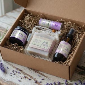 May include: A gift box filled with natural skincare products. The box contains a jar of balm, a bar of soap in a mesh bag, a lip balm, and a spray bottle. The products have lavender-themed labels and are nestled in brown paper filler.