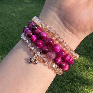 Puede incluir: Varios brazaletes de cuentas en tonos magenta y cristal transparente. Un brazalete tiene un dije de lazo dorado. Los brazaletes se muestran en una muñeca, sobre un fondo de hierba verde.