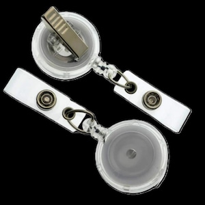 May include: Two clear plastic retractable badge holders with metal clips and snap-on straps. The badge reels have a clear, circular design with a metal clip on top and a snap-on strap at the bottom. The background is black.