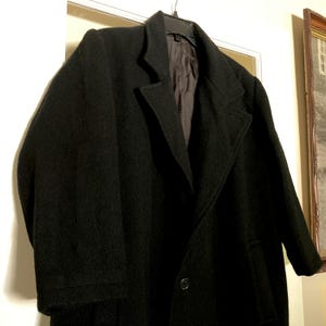 May include: A black wool-blend overcoat featuring a notched lapel, long sleeves, and a single-button front closure. The coat is fully lined with a dark interior fabric and is displayed on a black plastic hanger against a neutral background.