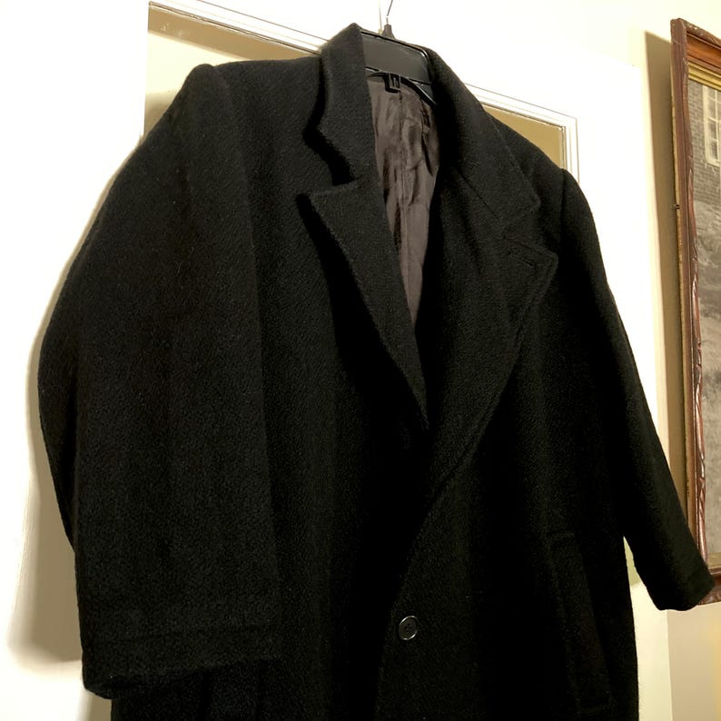 May include: A black wool-blend overcoat featuring a notched lapel, long sleeves, and a single-button front closure. The coat is fully lined with a dark interior fabric and is displayed on a black plastic hanger against a neutral background.