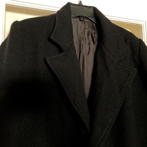 May include: A black coat with a textured surface, featuring wide lapels and a dark interior lining. The garment is displayed on a black plastic hanger, showcasing the structured collar design and the deep, solid black color of the outer fabric.