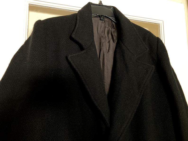 May include: A black coat with a textured surface, featuring wide lapels and a dark interior lining. The garment is displayed on a black plastic hanger, showcasing the structured collar design and the deep, solid black color of the outer fabric.