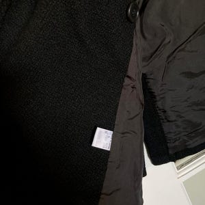 May include: A black textured coat featuring a smooth black lining and two dark buttons on the front. A white care label is sewn into the interior seam, displaying text in Japanese characters and indicating the garment was made in Japan.