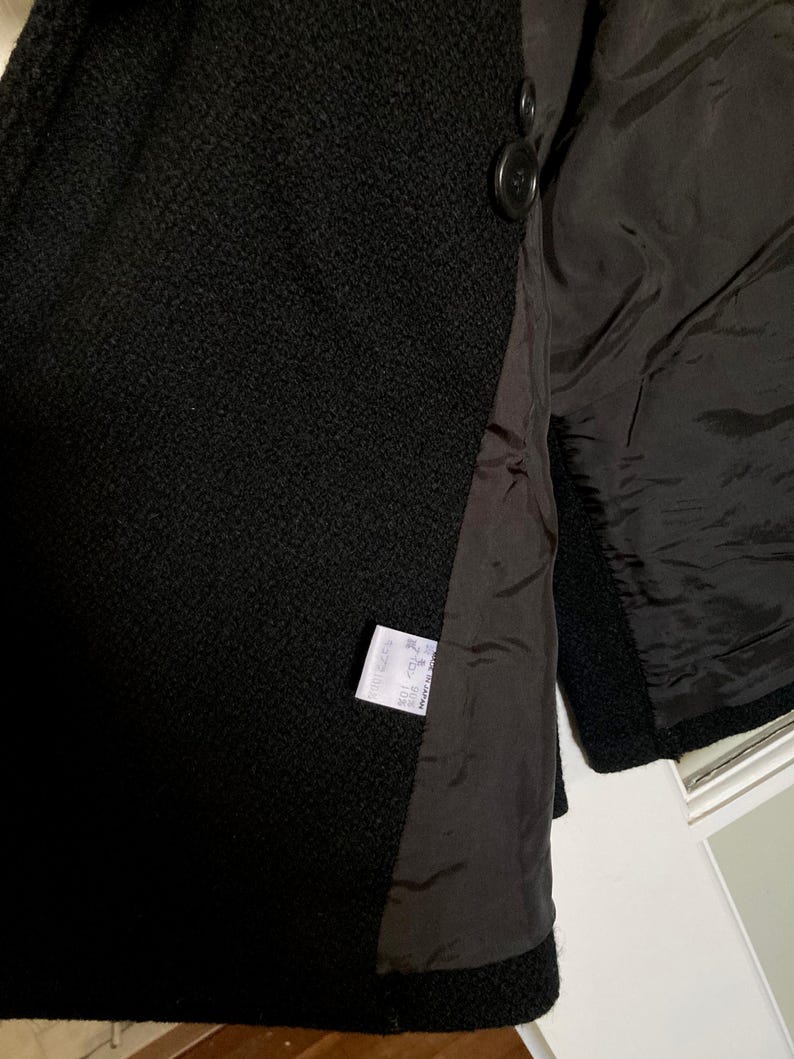 May include: A black textured coat featuring a smooth black lining and two dark buttons on the front. A white care label is sewn into the interior seam, displaying text in Japanese characters and indicating the garment was made in Japan.