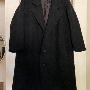 May include: A black wool-blend overcoat featuring a notched lapel, long sleeves, and a three-button front closure. The coat has a straight hemline and a dark brown interior lining, presented on a black plastic hanger against a white door.