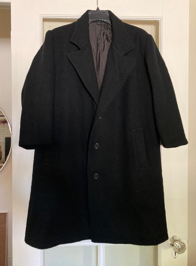 May include: A black wool-blend overcoat featuring a notched lapel, long sleeves, and a three-button front closure. The coat has a straight hemline and a dark brown interior lining, presented on a black plastic hanger against a white door.